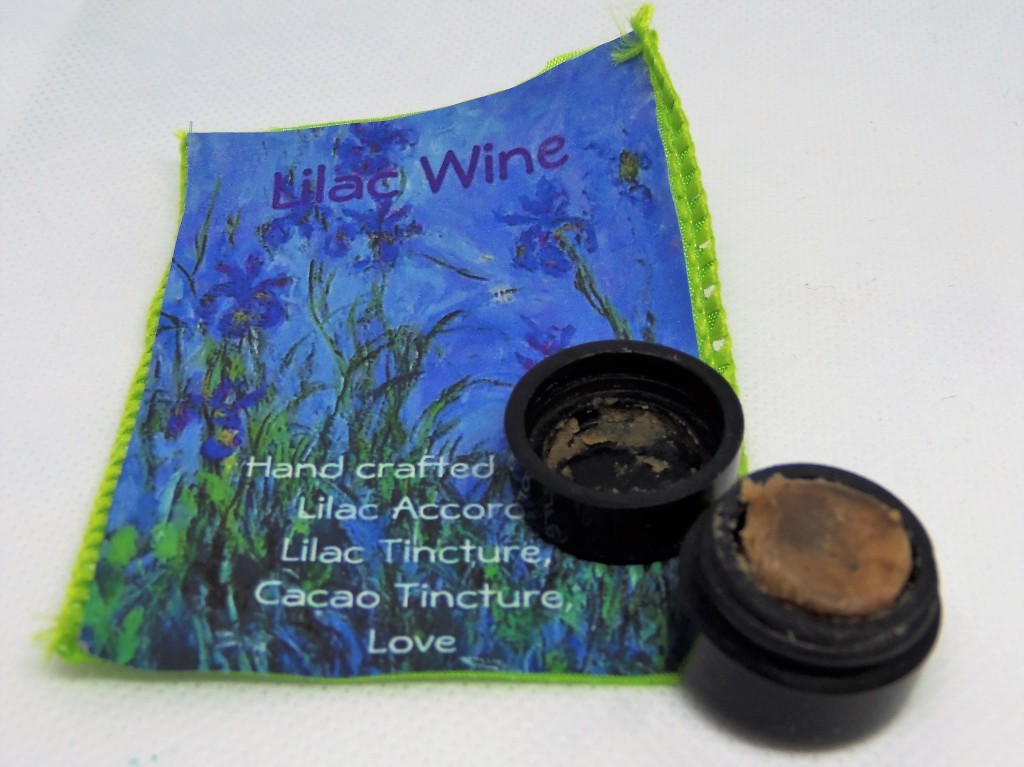 The Lilac Wine perfume, opened to show its dark brown color and paste-like texture, next to a printed card featuring illustrations of lilacs and a list of the notes:  lilac accord, lilac tincture, cacao tincture and love.