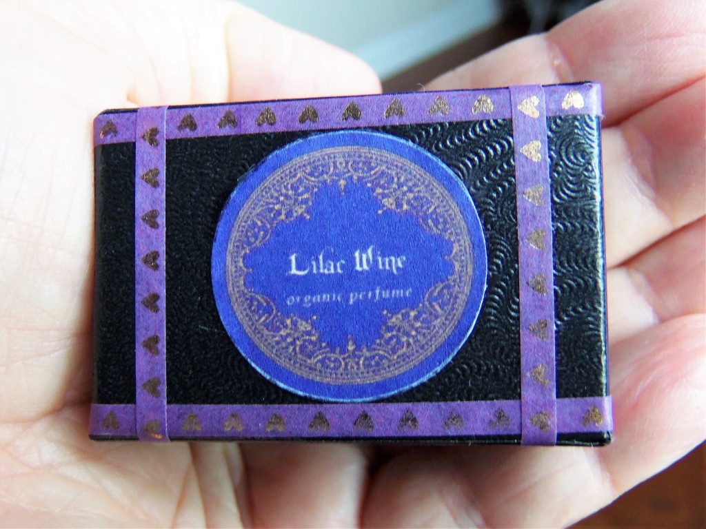 The black box for Lilac Wine perfume, with a purple label and purple decorative stripes stamped with tiny gold hearts